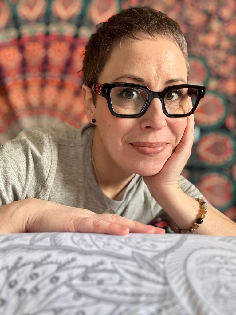 A portrait photograph of Erin, owner of Guided By Reiki, wearing black frame glasses and a casual grey t-shirt